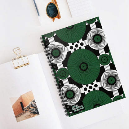 A bold green, black, and white African-inspired spiral notebook with geometric patterns lies on a white desk. Nearby are an open book with a wall photo, a gold binder clip, tortoiseshell sunglasses, and a white notepad.