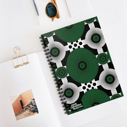 A bold green, black, and white African-inspired spiral notebook with geometric patterns lies on a white desk. Nearby are an open book with a wall photo, a gold binder clip, tortoiseshell sunglasses, and a white notepad.