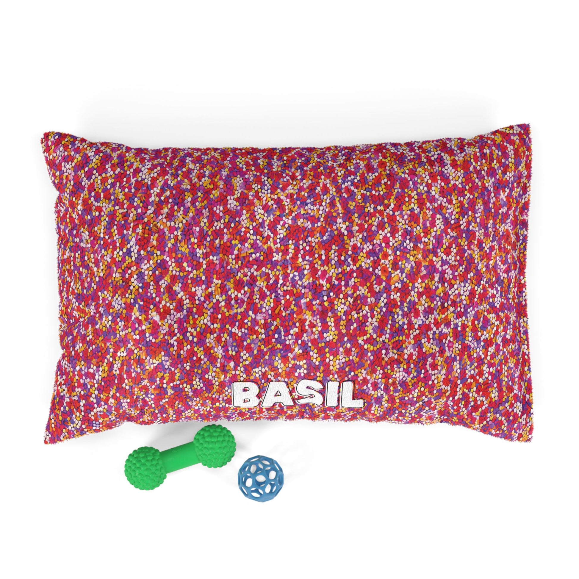 A rectangular, personalized pet bed with a colorful speckled pattern in red, pink, yellow, and white. BASIL is printed boldly on durable construction. In front are a green dumbbell-shaped toy and a small blue lattice ball.