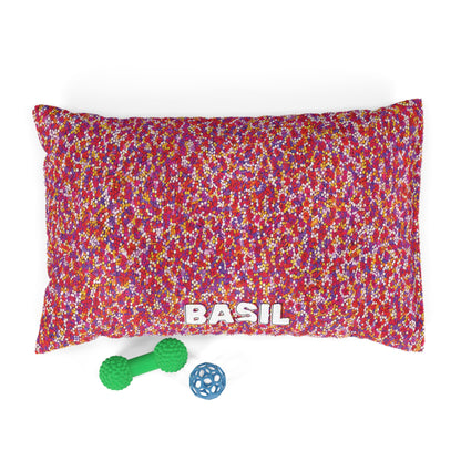 A rectangular, personalized pet bed with a colorful speckled pattern in red, pink, yellow, and white. BASIL is printed boldly on durable construction. In front are a green dumbbell-shaped toy and a small blue lattice ball.
