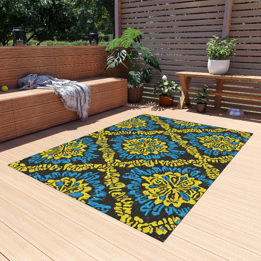 A modern outdoor patio with a tan wooden deck, wicker sofa draped with a gray blanket, green plants, and a wooden bench. Centered is an African-inspired outdoor rug featuring bold blue, yellow, and black floral patterns under sunlight filtering through wooden slats.