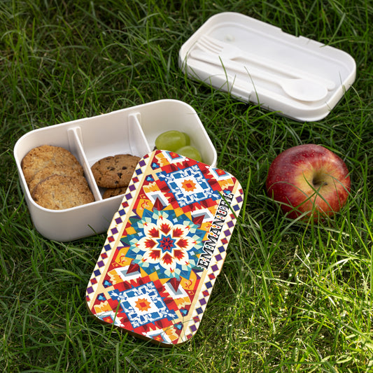 A personalized bento lunch box with a colorful geometric lid reading “EMMANUEL” sits on grass. The BPA-free lunchbox holds grapes and cookies in dividers, with a utensil compartment and a red apple nearby.