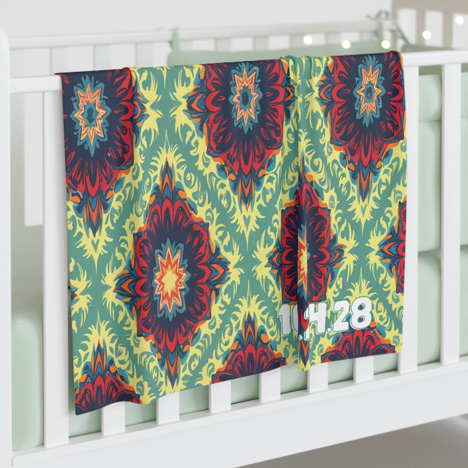 A colorful, African-inspired decor swaddle blanket with red, yellow, blue, and green mandala-like designs hangs over a white crib rail. The numbers 14:28 are printed in white near the bottom right corner. The crib bedding is light green.