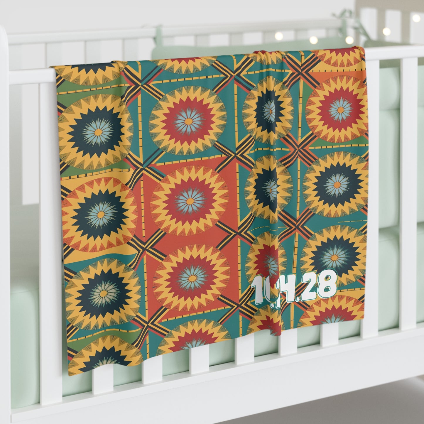 A colorful, African-inspired baby blanket with geometric, starburst-like designs in red, yellow, green, and blue hangs over a white crib rail. The numbers 1,14,28 in white personalize the blanket’s lower right side. Pale green bedding is inside the crib.