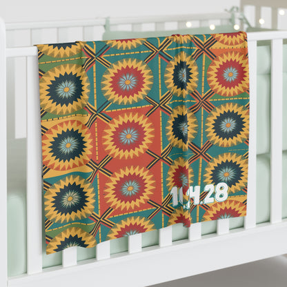 A colorful, African-inspired baby blanket with geometric, starburst-like designs in red, yellow, green, and blue hangs over a white crib rail. The numbers 1,14,28 in white personalize the blanket’s lower right side. Pale green bedding is inside the crib.