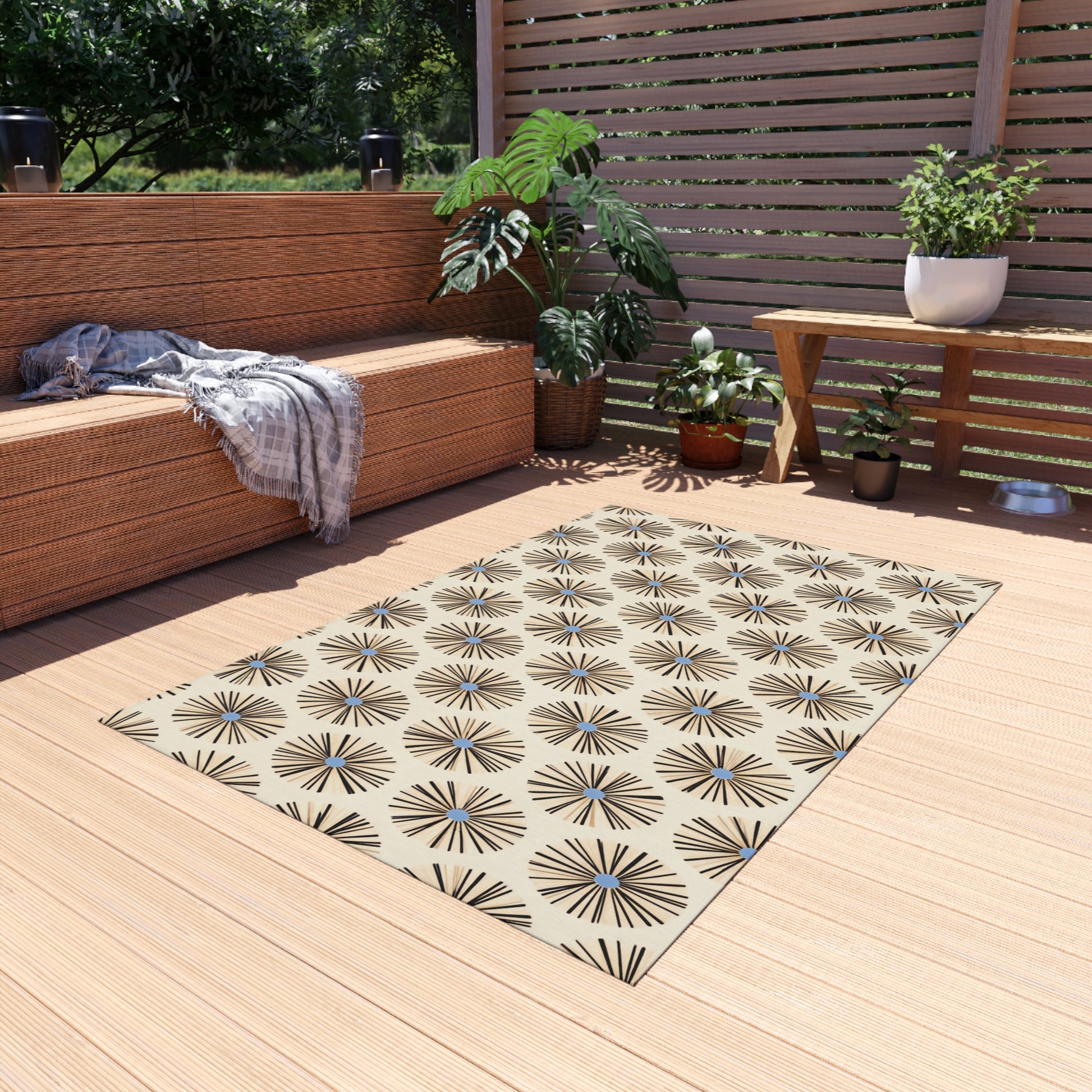 Outdoor patio deck with a woven couch, gray striped blanket, wooden bench, and potted plants. A beige African-inspired outdoor rug with a geometric starburst pattern lies on light wood flooring. Sunlight filters through slatted fencing and trees.