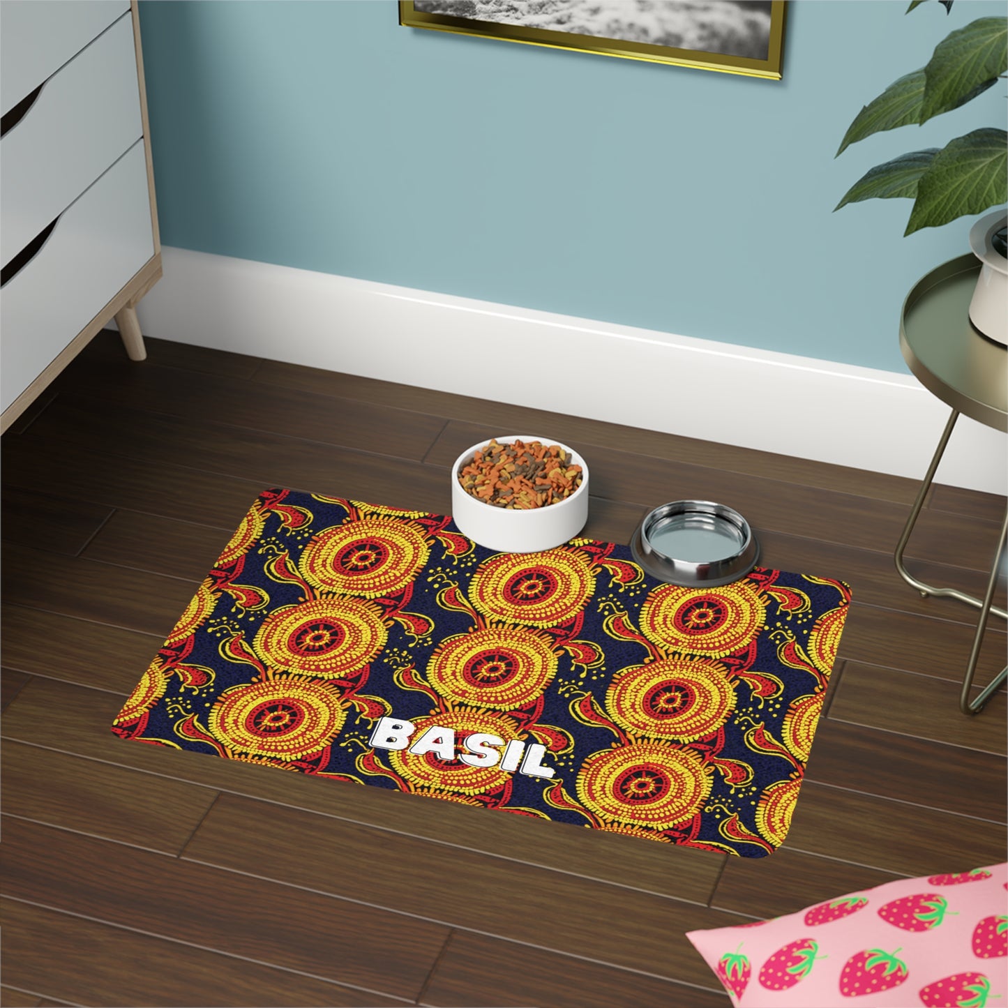 A pet feeding station on a dark wood floor features an African-inspired mat with bright yellow and red circular patterns and the name BASIL in bold white letters. On the mat are a bowl of dry pet food, a metal water dish, a plant, and a nearby drawer.