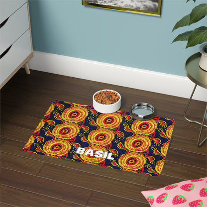 A pet feeding station on a dark wood floor features an African-inspired mat with bright yellow and red circular patterns and the name BASIL in bold white letters. On the mat are a bowl of dry pet food, a metal water dish, a plant, and a nearby drawer.