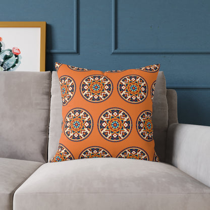 A square orange African-inspired pillow with circular, mosaic-style geometric patterns in blue, white, red, and yellow rests on a light gray sofa against a dark blue paneled wall, with framed cactus artwork partially visible to the left.