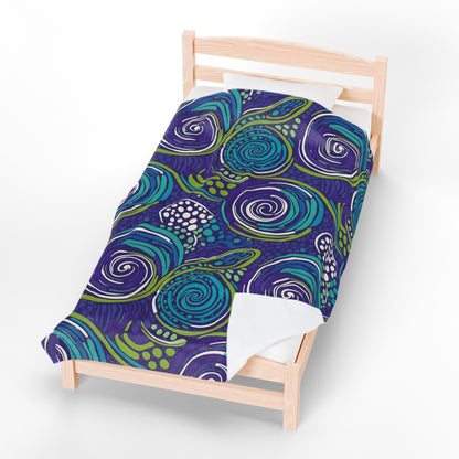 A single wooden bed with a personalized velveteen blanket featuring a bold abstract design of swirling blue, teal, and green patterns with white accents on a dark purple background. The bed has a white pillow and is photographed on a plain white surface.