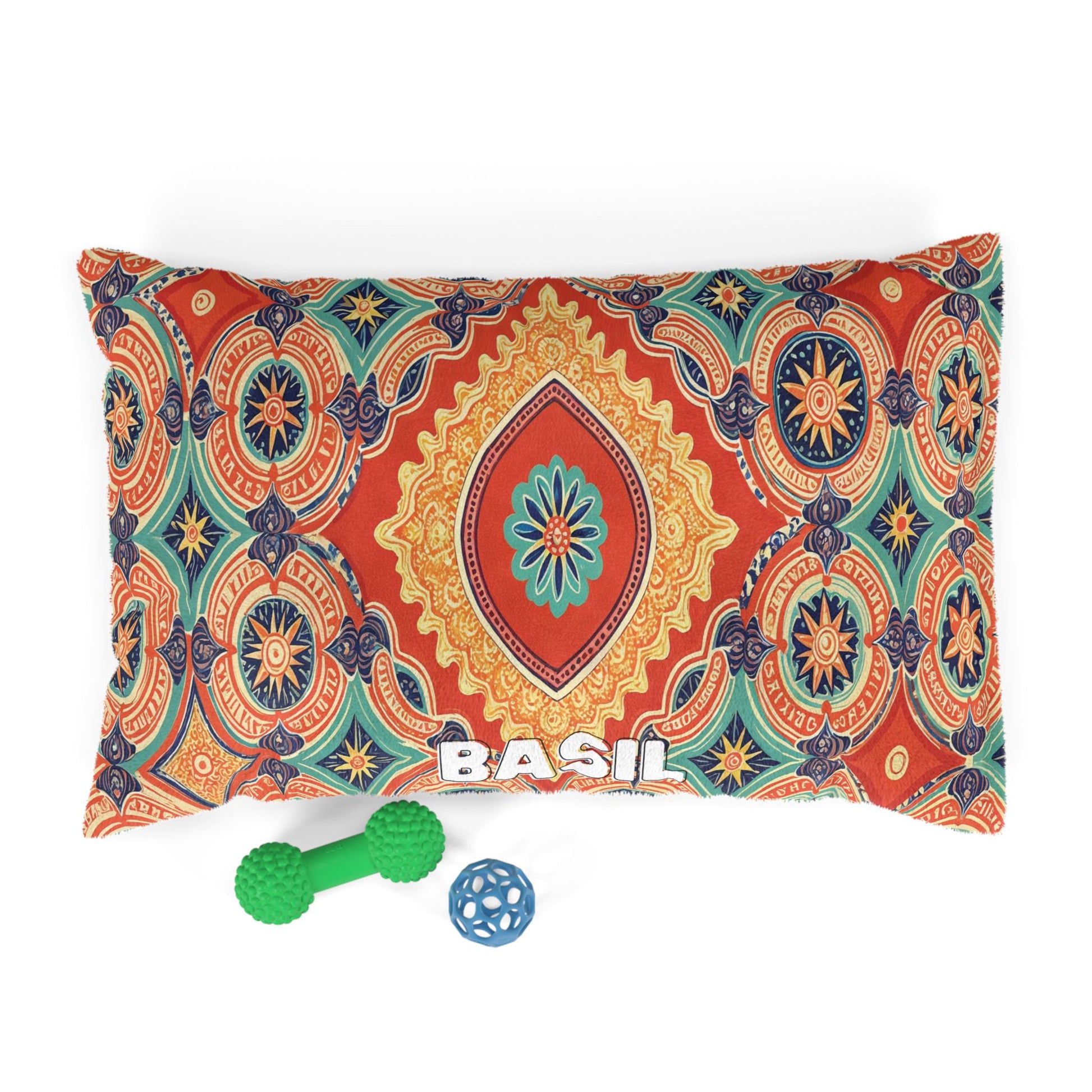 A custom pet pillow in a rectangular shape displays an intricate orange, blue, and gold mandala pattern with BASIL in white letters. In front sit two green textured dog toys and a small blue ball with holes—a perfect accent for a stylish dog bed.