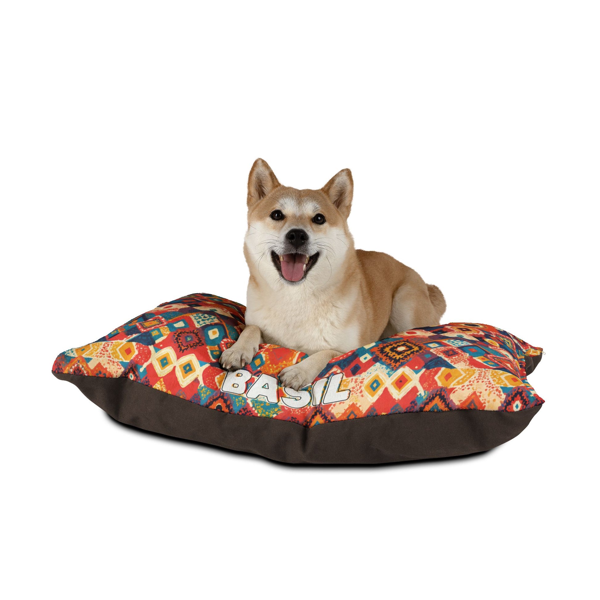 A happy Shiba Inu dog with tan fur lies on a colorful African-inspired patterned pillow with “BASIL” written on it. The dog’s mouth is open, tongue out, and ears perked up. The plain white background highlights the dog and pillow.