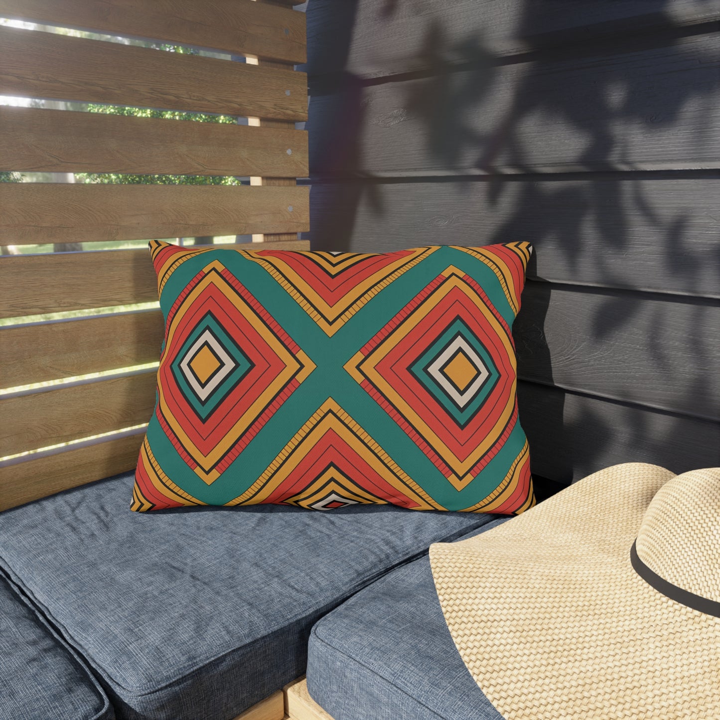 A square outdoor pillow with a bold geometric pattern in red, orange, yellow, green, and black rests on a blue cushioned bench. Made from weatherproof materials, it sits beside a woven straw hat as light and shadows dance across the scene.