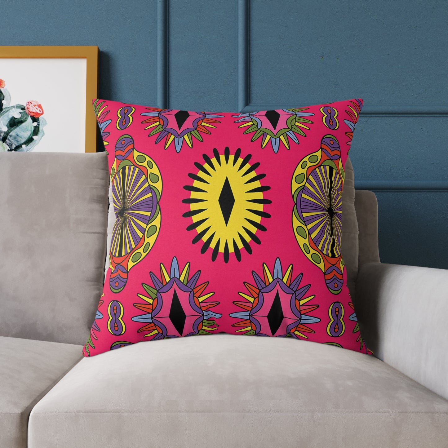 A vibrant home decor touch, this colorful accent pillow features bold geometric patterns in pink, yellow, orange, and black. It sits on a light gray sofa against a deep blue wall, with a framed cactus art piece in the background.