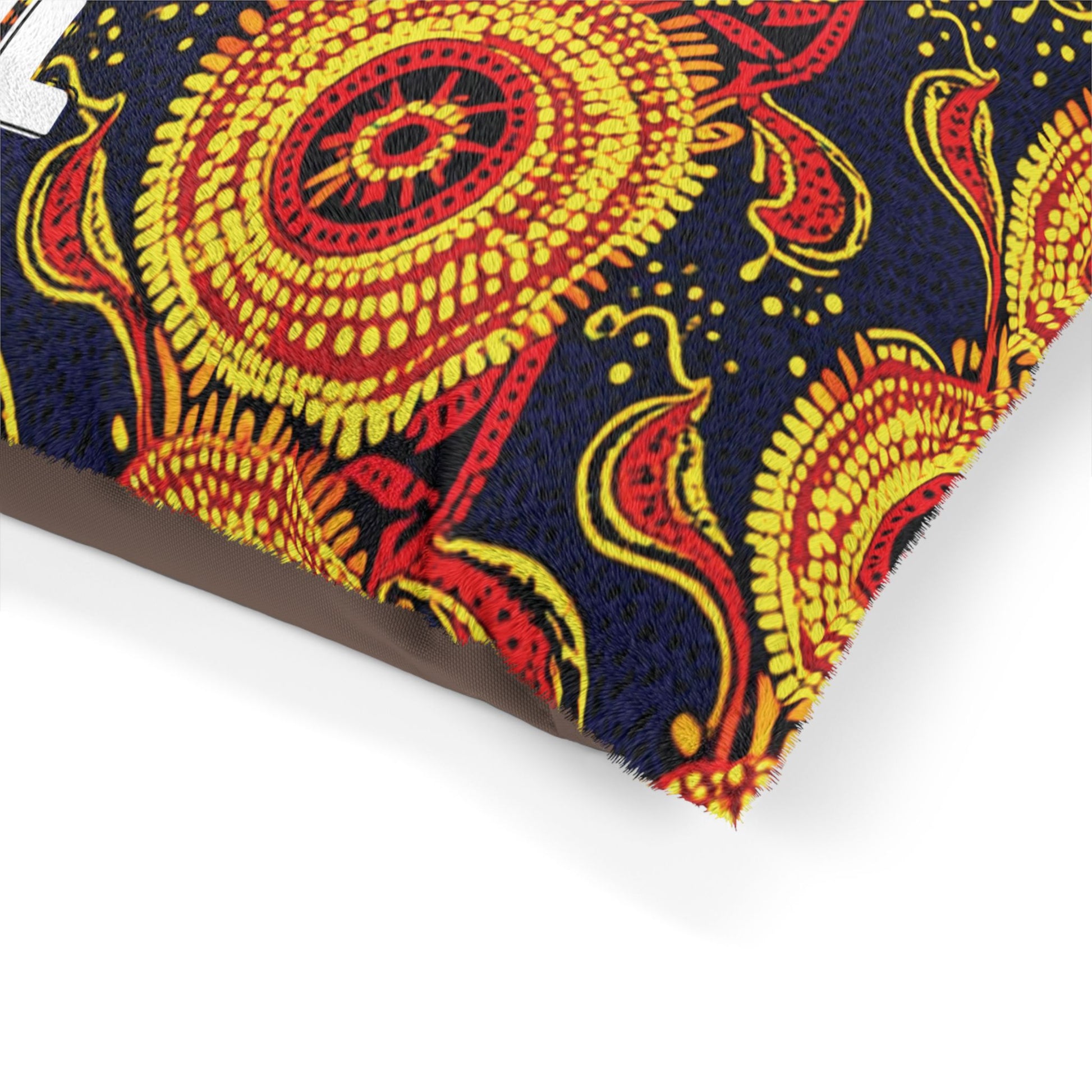 A close-up of a decorative African-inspired fabric pillow featuring bold, intricate patterns in bright yellow and red on a dark blue background, with swirling floral and circular motifs and a small corner of a white letter partially visible on the left side.