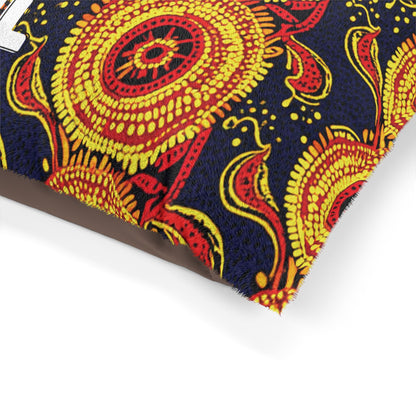 A close-up of a decorative African-inspired fabric pillow featuring bold, intricate patterns in bright yellow and red on a dark blue background, with swirling floral and circular motifs and a small corner of a white letter partially visible on the left side.