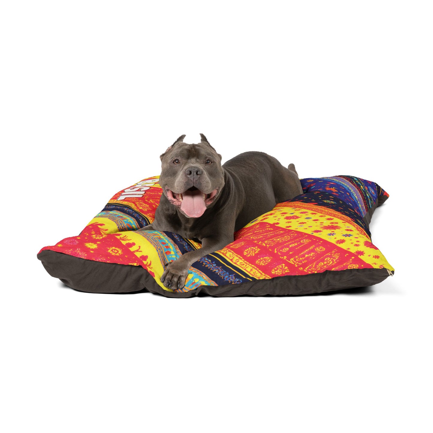A gray pit bull with cropped ears lies happily on a large, African-inspired dog bed with red, yellow, and blue patterns. The dog is panting, tongue out and front paws extended, looking content against a white background.