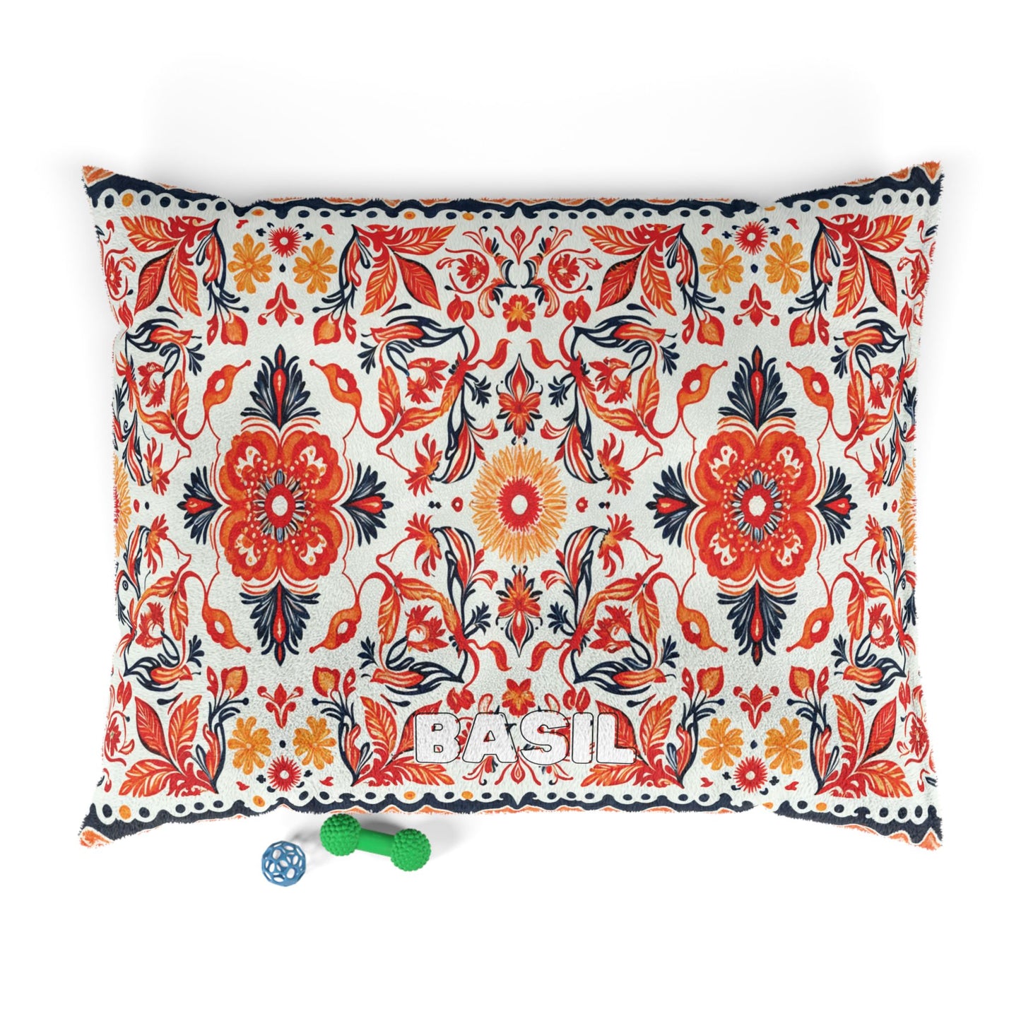 Rectangular pillow with a vibrant red, orange, and navy African-inspired floral pattern. The name “BASIL” is stitched in white at the bottom center. Below are a green bone-shaped chew toy and a blue ball with white designs on a white background.