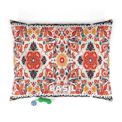 Rectangular pillow with a vibrant red, orange, and navy African-inspired floral pattern. The name “BASIL” is stitched in white at the bottom center. Below are a green bone-shaped chew toy and a blue ball with white designs on a white background.