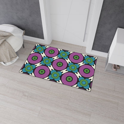 Rectangular rug on light wood floor features a bold, African-inspired geometric pattern with concentric circles in purple, turquoise, and green, separated by black lines on a cream background. The rug is near a white cabinet and beige throw blanket.