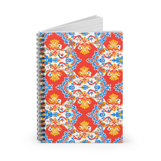 A spiral-bound notebook with a vibrant cover featuring a symmetrical red, blue, and yellow floral-inspired pattern on a white background draws from culturally inspired designs. The notebook is open slightly, revealing white, lined pages inside.