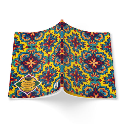 An open African-inspired journal features a vibrant, symmetrical floral pattern in blue, red, black, and orange on a yellow background. A gold hexagonal label on the back cover reads AFRO COMFORT SHOP with contact details below. Viewed from above.