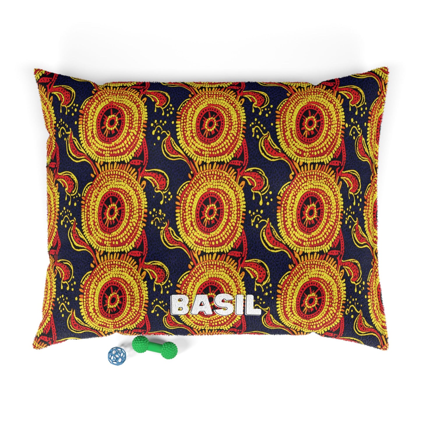 A rectangular pet bed with a bold African-inspired red, yellow, and black geometric pattern is seen from above. BASIL is printed in large white letters on the front. Below the bed are two dog toys: a green bone with texture and a blue ball.