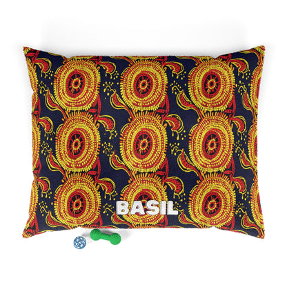 A rectangular pet bed with a bold African-inspired red, yellow, and black geometric pattern is seen from above. BASIL is printed in large white letters on the front. Below the bed are two dog toys: a green bone with texture and a blue ball.