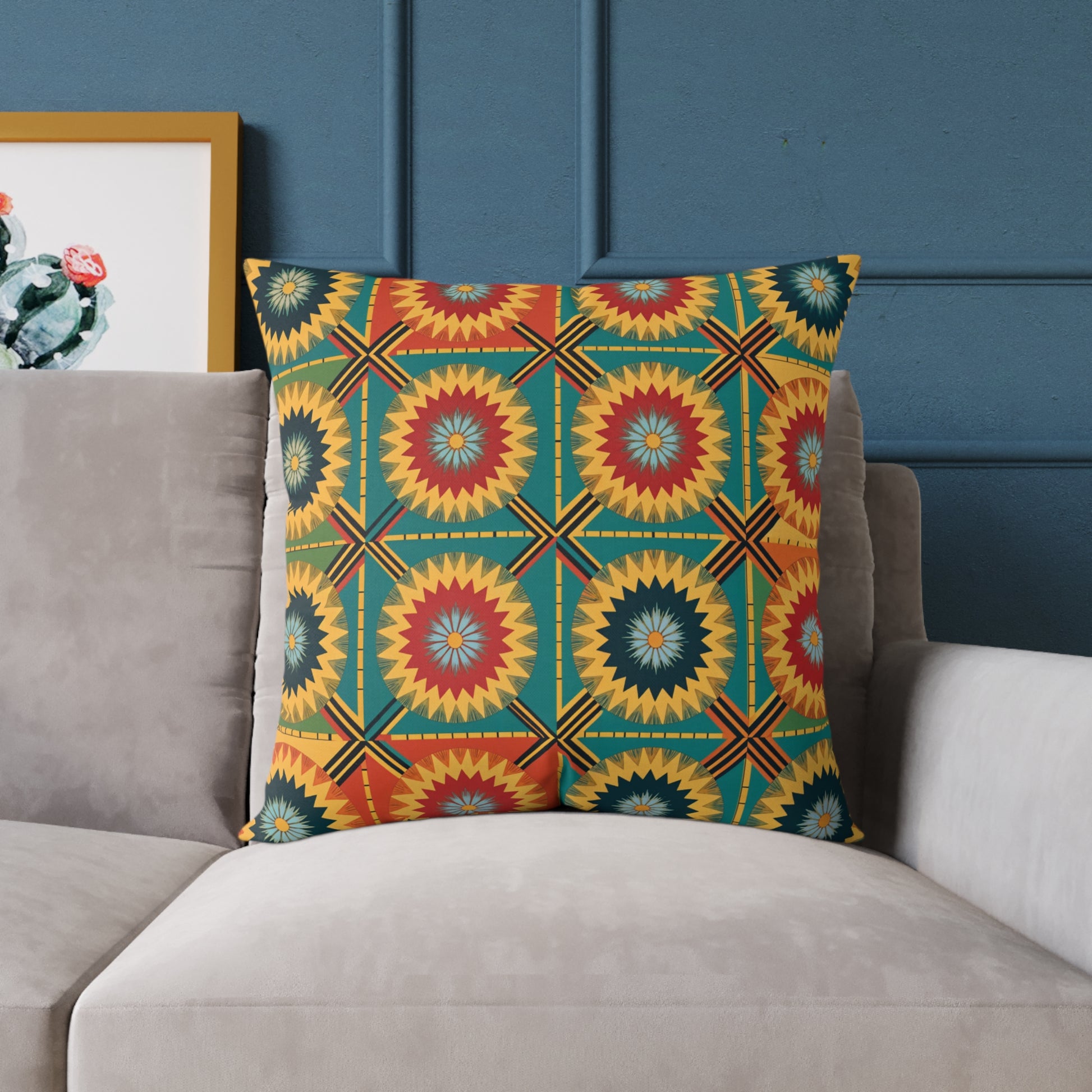 A colorful accent pillow with a bold geometric pattern in red, yellow, teal, and blue sits on a light gray couch. The wall behind is dark blue, and a framed artwork with a cactus design is partially visible on the left.