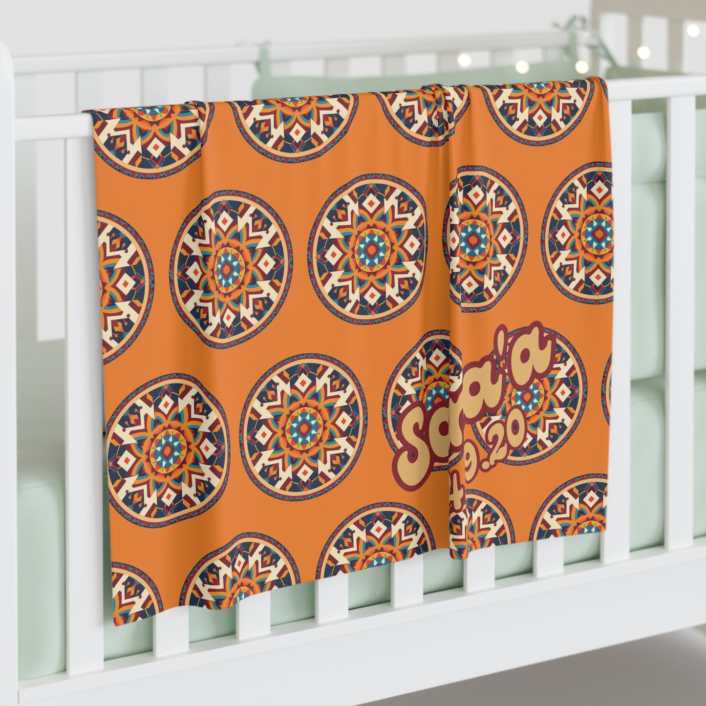 A soft orange personalized baby blanket with colorful, circular geometric patterns and the text Safaa 2020 draped over a white crib. Light mint green bedding and string lights add charm to this African-inspired decor nursery.