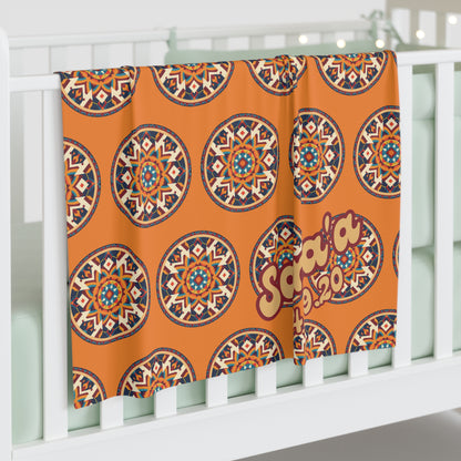 A soft orange personalized baby blanket with colorful, circular geometric patterns and the text Safaa 2020 draped over a white crib. Light mint green bedding and string lights add charm to this African-inspired decor nursery.
