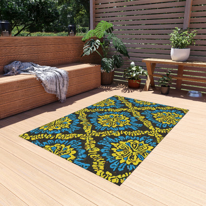 A cozy outdoor patio with light wooden decking features a wicker sofa topped with gray cushions and a plaid blanket. An African-inspired outdoor rug in blue and yellow patterns brightens the floor, while potted plants and a slat screen complete the relaxing scene.