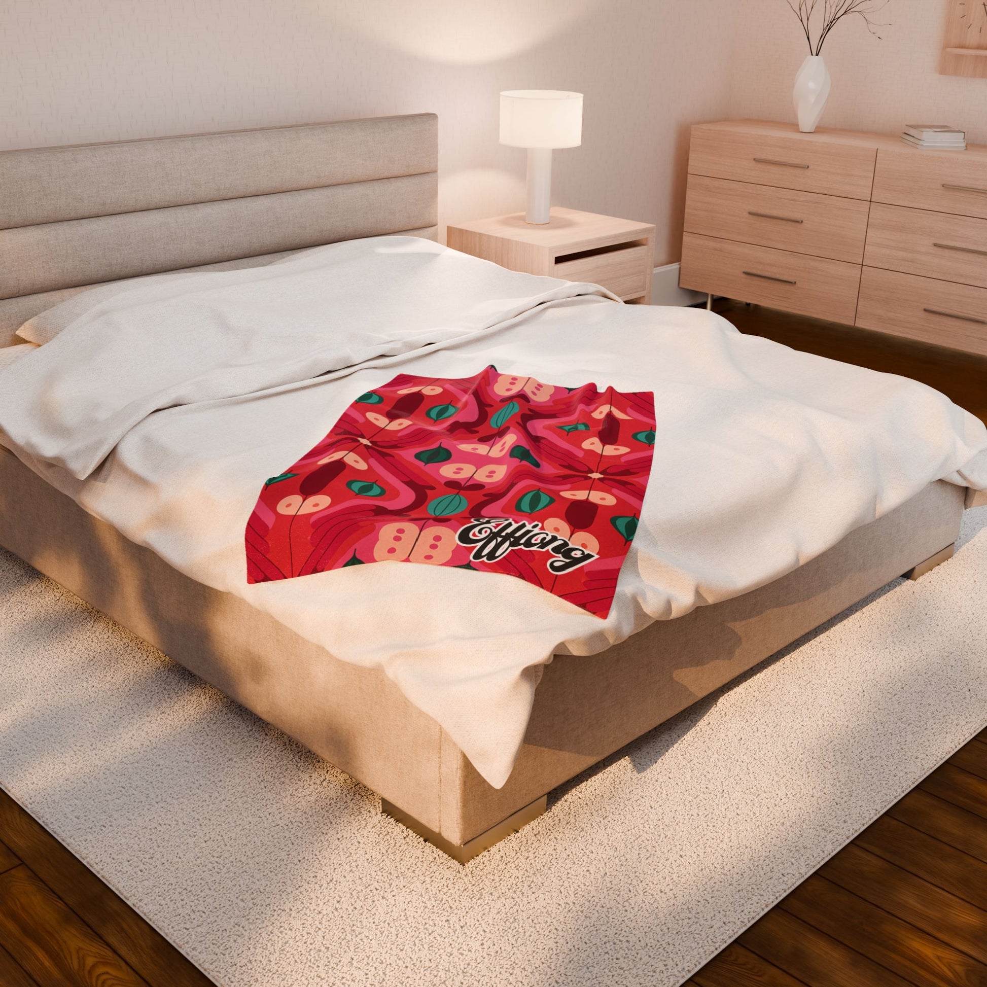 A beige bed with white bedding is topped by a personalized velveteen blanket featuring colorful abstract apples and the word “Apples” in script. The room showcases wooden floors, light wood furniture, and sunlit charm.