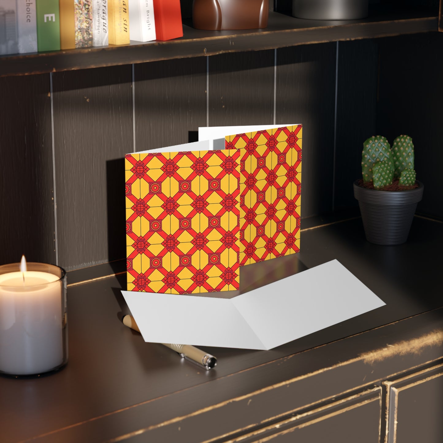 A geometric-patterned greeting card with African-inspired designs in red and yellow stands open on a dark shelf. Nearby are a blank white card, a pen, a small potted cactus, a lit candle, and several books. Warm lighting creates a cozy atmosphere.