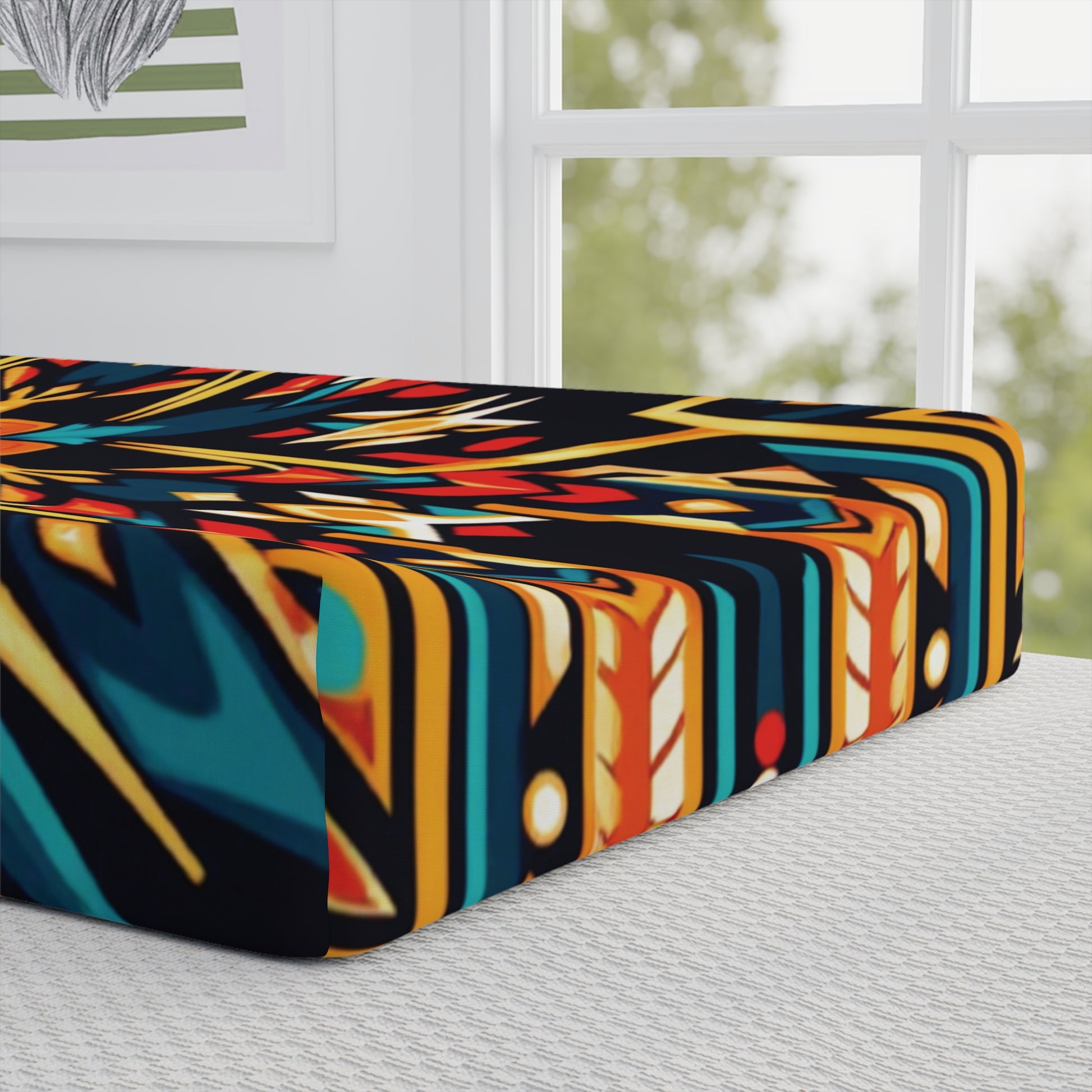A close-up of a mattress or cushion with a vibrant geometric pattern in blue, yellow, red, and black showcases African-inspired designs. The background includes a large window with trees outside and framed wall art with a leaf motif. The bed has a textured white cover.