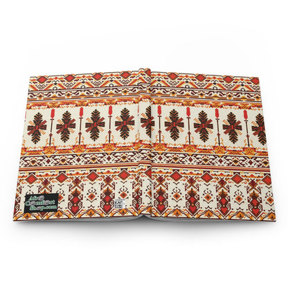 A hardcover journal with a cover featuring an intricate, symmetrical, tribal pattern notebook design in red, brown, gold, and cream. Rows of stylized leaves and geometric shapes adorn it, with a small QR code and green label in the lower left corner.