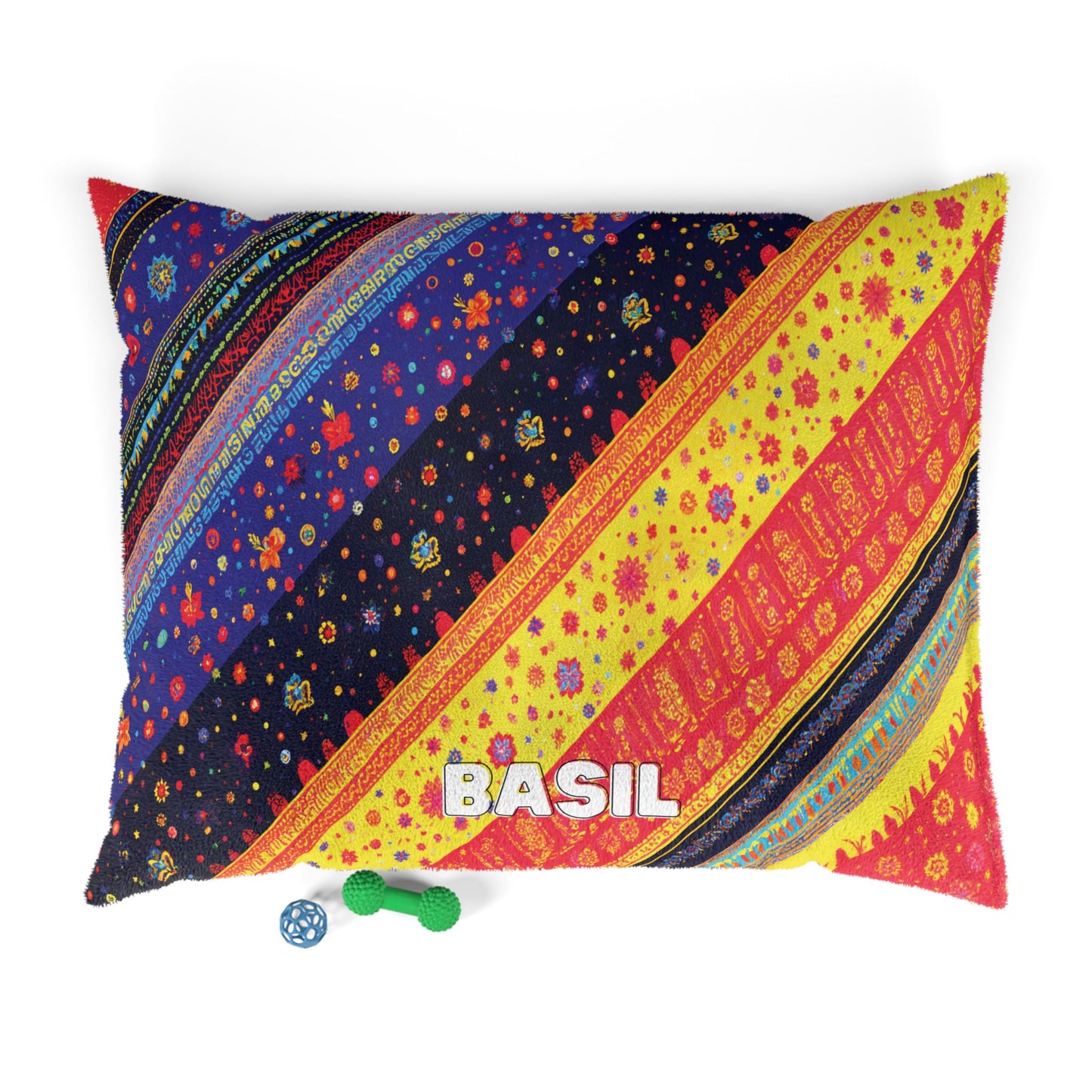 A colorful, African-inspired rectangular pillow with diagonal stripes in blue, yellow, red, and orange patterns features the name BASIL in white letters. Next to the pillow are a green textured bone toy and a blue rubber ball with holes.
