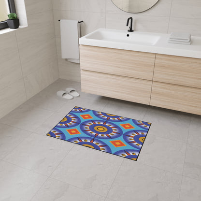 A modern bathroom with light wood vanity, white sink, black faucet, and round mirror. Custom floor mats with colorful geometric patterns and non-slip rubber backing add African-inspired aesthetics to the tiled floor. White slippers rest near a window.