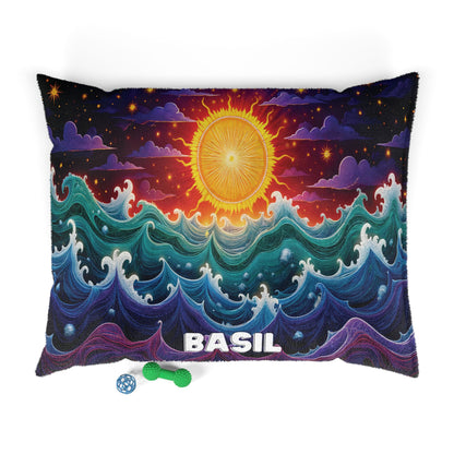 A rectangular personalized pet bed with the name BASIL at the bottom shows a colorful, stylized ocean with swirling waves and a radiant yellow sun above, set against a starry, partly cloudy sky. Below are a green textured bone toy and a blue ball.