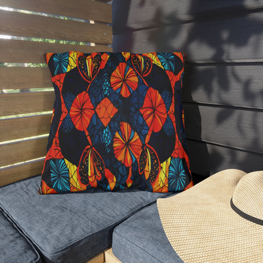 A square, UV-resistant outdoor pillow with a colorful butterfly wing pattern in orange, blue, yellow, and black rests on a blue water-resistant cushion. Next to it sits a straw hat in a shaded patio corner with wooden slats and leafy shadows.