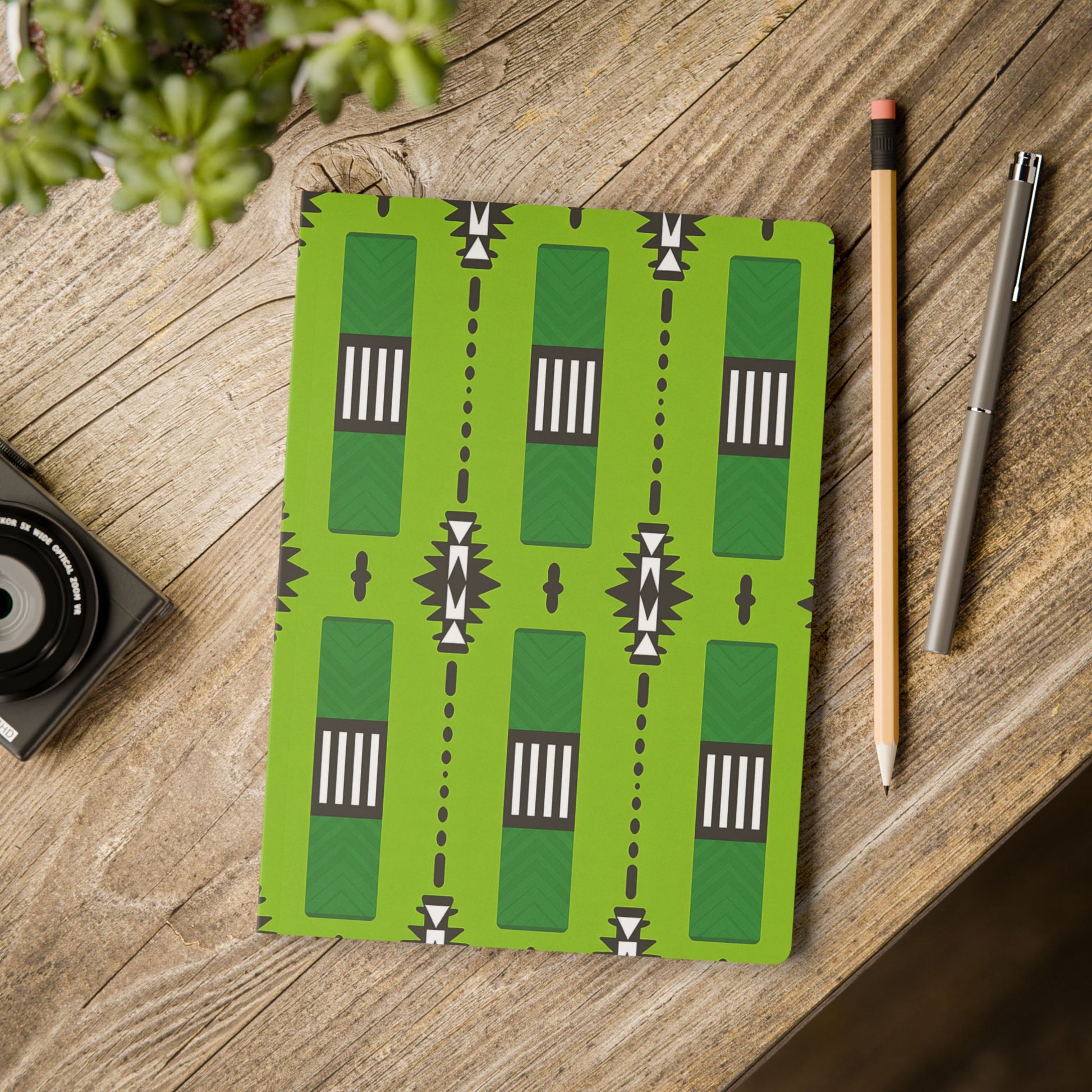 A green African-inspired journal with a bold, geometric pattern in black, white, and dark green lies on a rustic wooden table—perfect for daily journaling. Beside it are a pencil, silver pen, camera, and a potted succulent in the corner.
