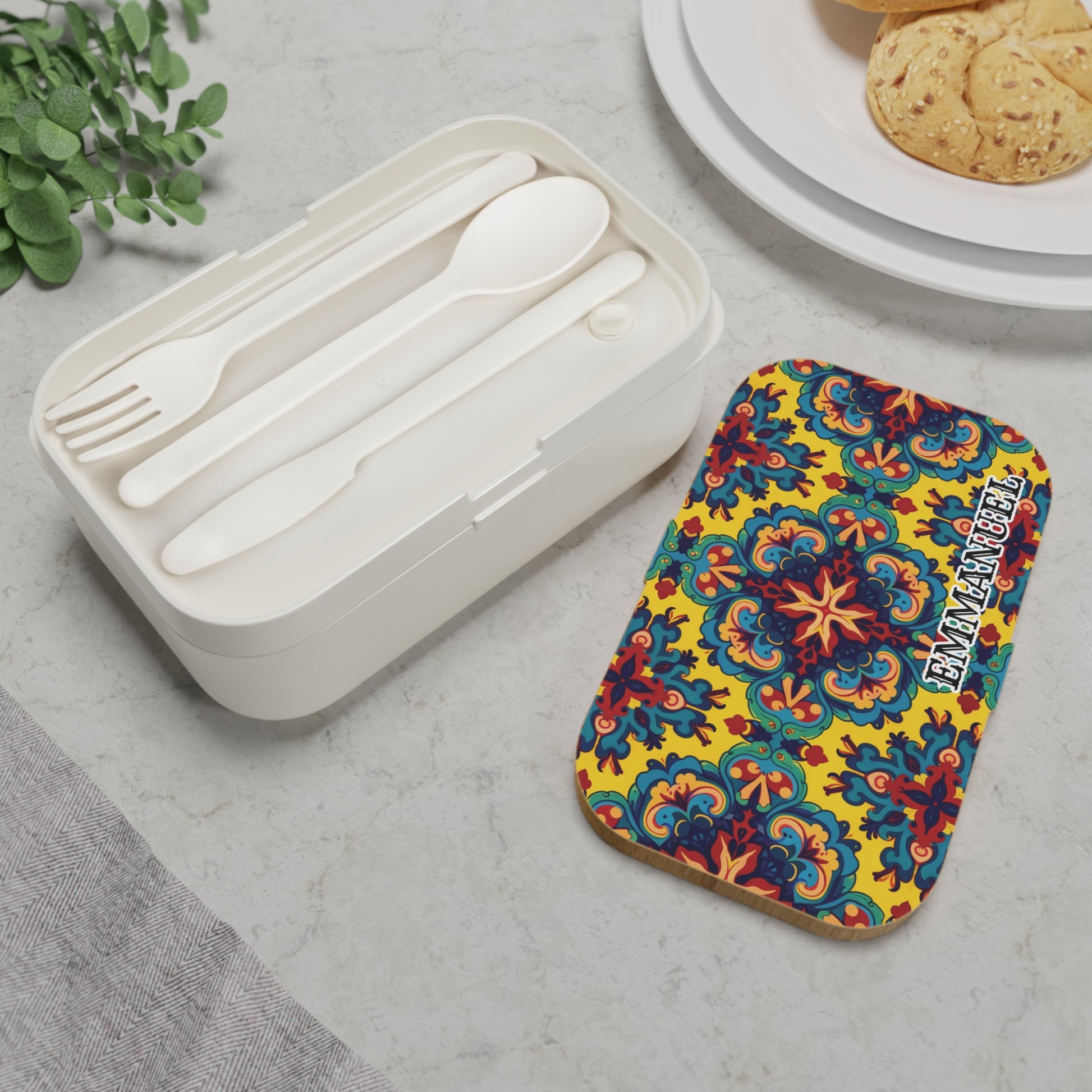 A personalized bento lunch box in white with matching cutlery sits beside a vibrant, floral SAN MIGUEL lid. The African-inspired lunch box rests on marble near a plant, gray napkin, and cookies for a stylish meal setup.