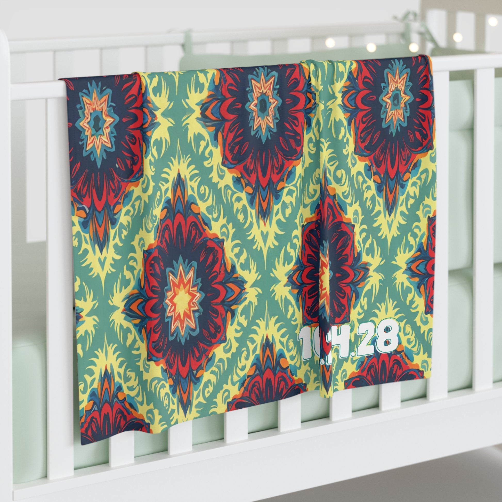 A colorful, personalized baby blanket with a bold, abstract floral pattern in red, yellow, blue, and green hangs over a white crib. The numbers 14 28 in white are printed near the corner. Perfect for African-inspired decor with pale green crib bedding.