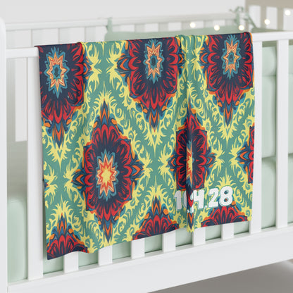 A colorful, personalized baby blanket with a bold, abstract floral pattern in red, yellow, blue, and green hangs over a white crib. The numbers 14 28 in white are printed near the corner. Perfect for African-inspired decor with pale green crib bedding.