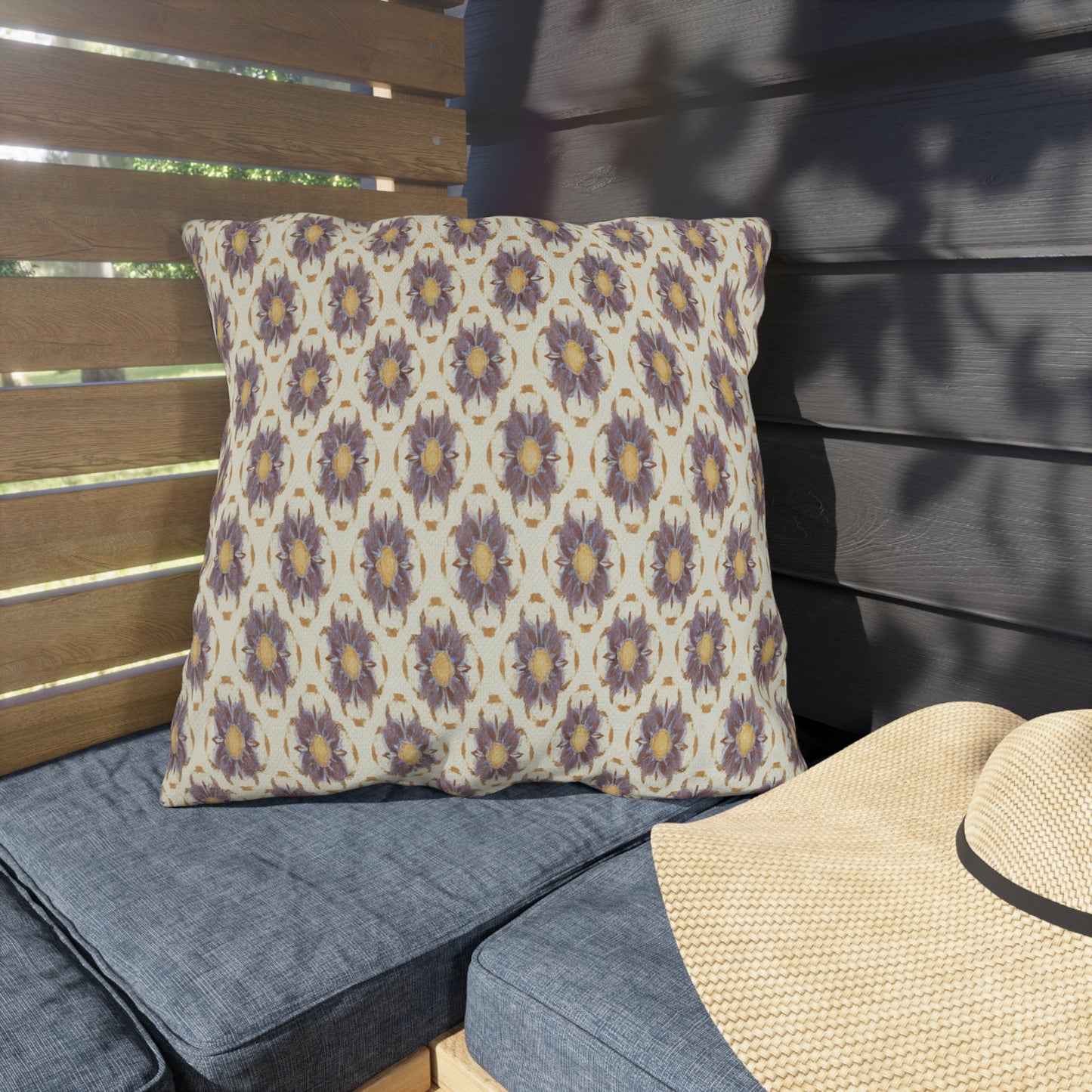 A UV-resistant outdoor pillow with a beige background and a repeating purple and yellow floral pattern rests on a blue cushioned bench. Next to it is a straw hat, while wooden slats cast gentle shadows across the cozy space.