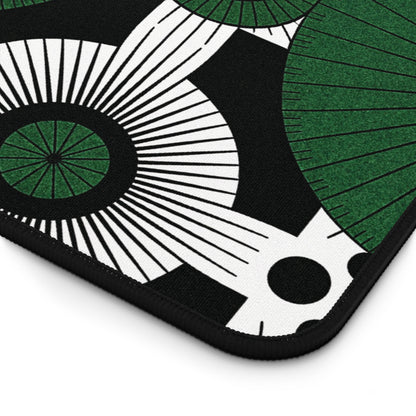 A close-up of an ergonomic desk pad featuring a modern, abstract pattern. Green and white circular shapes with radiating lines, black centers, and black polka dots stand out on the black background. The neoprene desk mat has rounded, black-stitched edges.