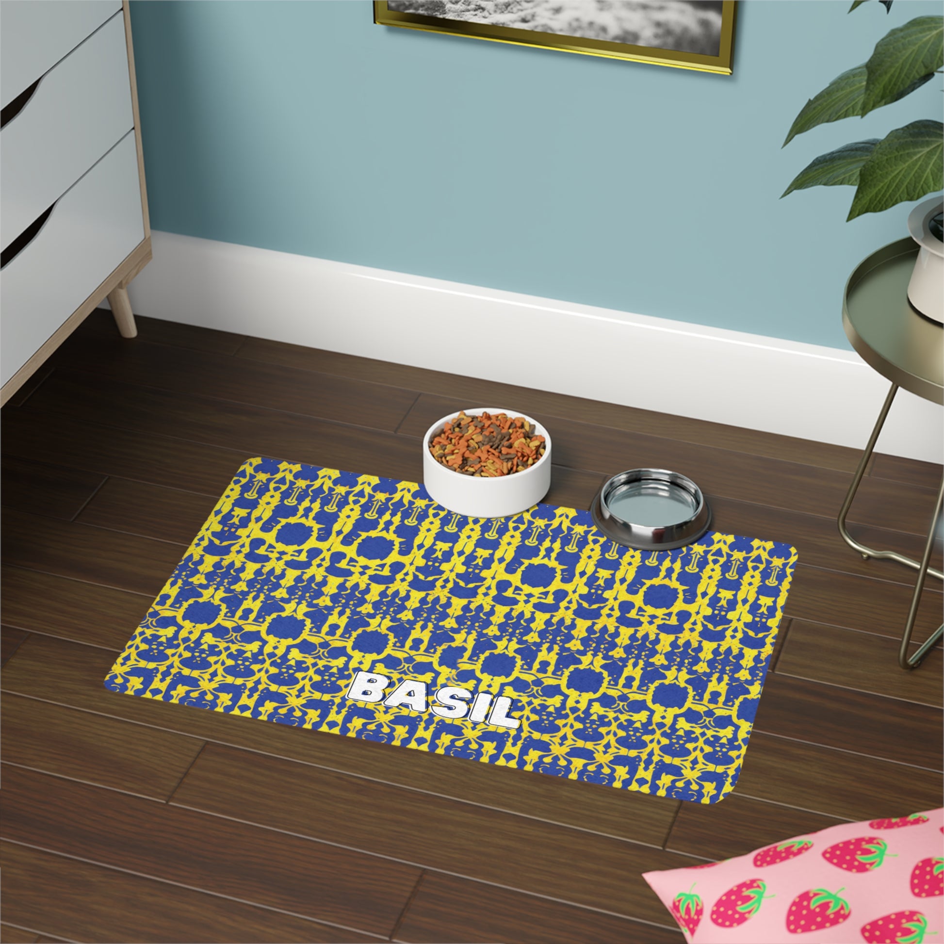 A patterned, African-inspired blue and yellow pet mat labeled “BASIL” sits on a dark wood floor. On the mat are a bowl of dry pet food and a stainless steel water bowl. Nearby are a dresser, green plant, pink strawberry mat, and framed picture on a blue wall.