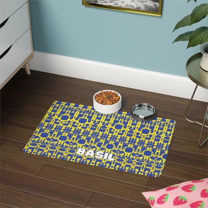 A patterned, African-inspired blue and yellow pet mat labeled “BASIL” sits on a dark wood floor. On the mat are a bowl of dry pet food and a stainless steel water bowl. Nearby are a dresser, green plant, pink strawberry mat, and framed picture on a blue wall.