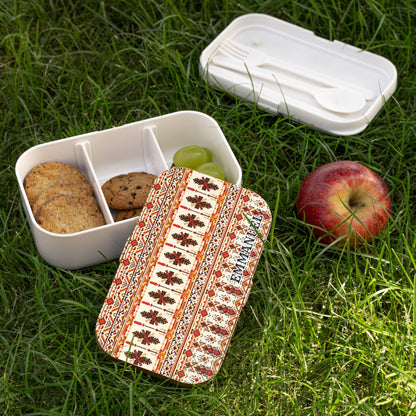 A patterned African-inspired lunch box sits on grass, lid off, revealing sections with cookies and green grapes. A red apple is nearby. The lid showcases a red and brown floral geometric design. A separate white compartment holds a spoon and fork.