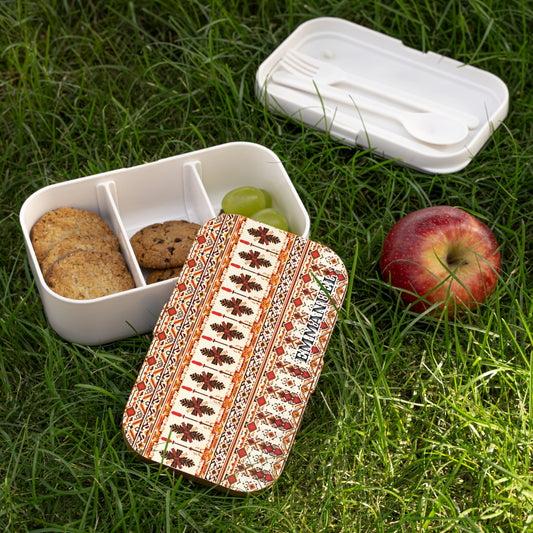 A patterned African-inspired lunch box sits on grass, lid off, revealing sections with cookies and green grapes. A red apple is nearby. The lid showcases a red and brown floral geometric design. A separate white compartment holds a spoon and fork.